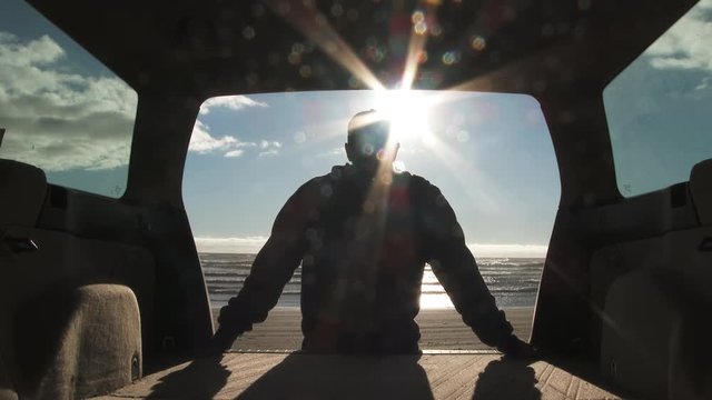 Silhouetted man sits down on back of car, relaxing at the beach while watching the sun go down over the Pacific Ocean in Oregon.