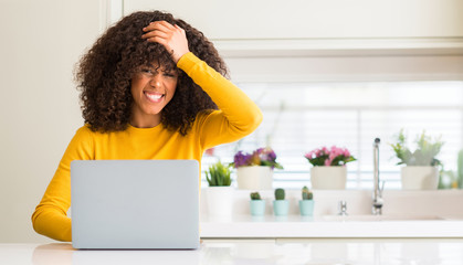 African american woman using computer laptop at kitchen stressed with hand on head, shocked with...
