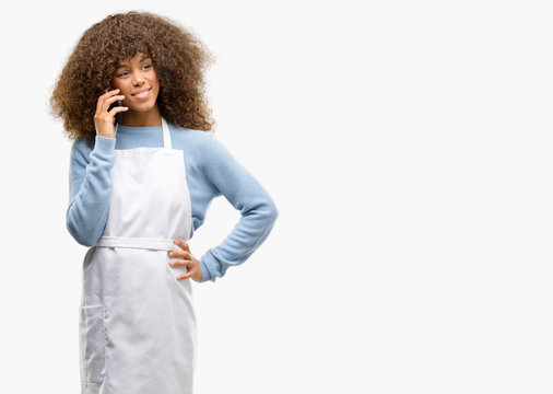 African american shop owner woman wearing an apron happy talking using a smartphone mobile phone