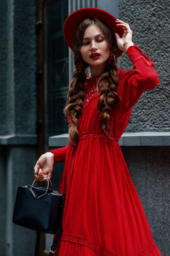 Outdoor portrait of young beautiful fashionable woman wearing total red look: dress, hat, earrings, holding black small learther bad, posing in street of the city. Female fashion concept

