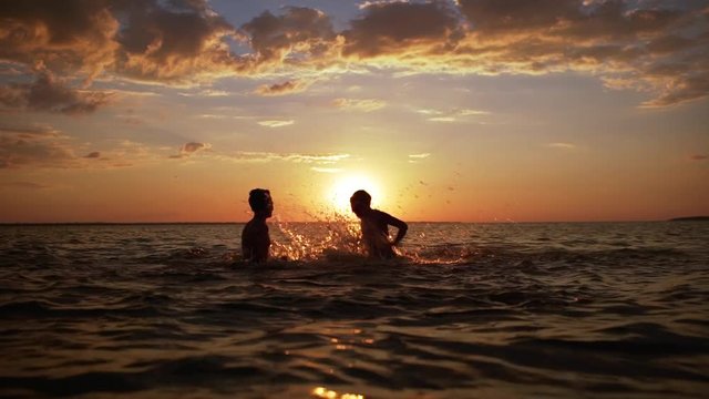 Silhouettes of two cheerful boys 10-12 having fun and splashing water together, while swimming in sea during beautiful sunset in slow motion