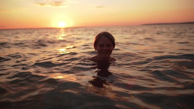 Portrait of young caucasian boy 10-12 swimming in sea alone and looking at you, during amazing dusk in slow motion