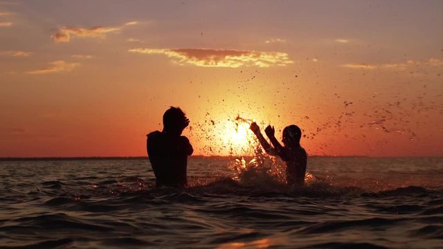 Silhouettes of adult father and little son fooling around and splashing water, while swimming in ocean during beautiful sunset in slow motion