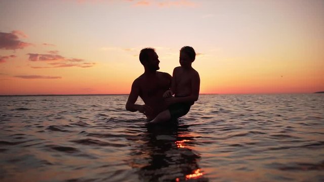 Silhouettes of happy father carrying his little son in arms in water, after swimming in ocean during beautiful sunset in slow motion