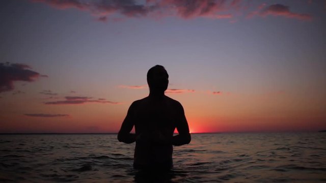 Silhouette of handsome adult man walking in water back to coast, after swimming in sea during beautiful sunset in slow motion