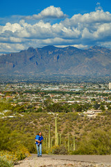 Fototapeta na wymiar Grandfather Carries His Young Grandson Up the Steep Paved Path of A Mountain in Tucson, Arizona With the City in the Background