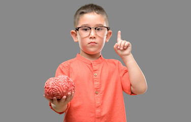 Dark haired little child with glasses holding a brain surprised with an idea or question pointing...