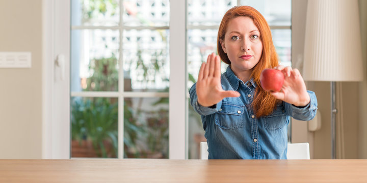 Redhead woman holding red apple at home with open hand doing stop sign with serious and confident expression, defense gesture