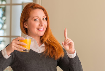 Redhead woman holding orange juice glass at home, surprised with an idea or question pointing finger with happy face, number one