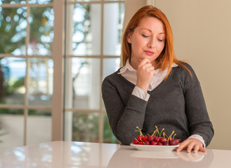 Redhead woman eating cherries at home serious face thinking about question, very confused idea