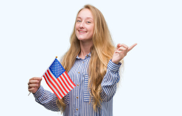 Blonde teenager woman holding flag of United States of America very happy pointing with hand and...