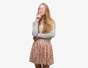 Blonde teenager woman wearing flowers skirt with hand on chin thinking about question, pensive expression. Smiling with thoughtful face. Doubt concept.