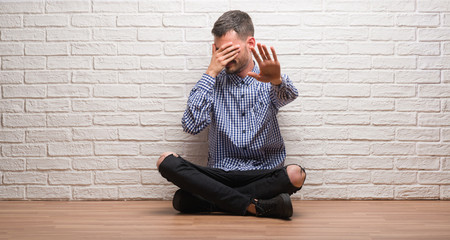 Young adult man sitting over white brick wall covering eyes with hands and doing stop gesture with sad and fear expression. Embarrassed and negative concept.