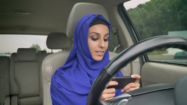 Young muslim woman in hijab sitting behind self-driving steering wheel of autonomous autopilot driverless car and typing on cell phone