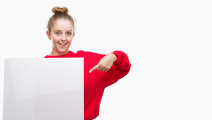 Young blonde woman holding advertising banner very happy pointing with hand and finger