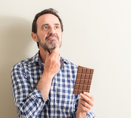Senior man eating chocolate bar serious face thinking about question, very confused idea