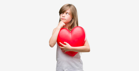 Young blonde toddler holding a red heart serious face thinking about question, very confused idea