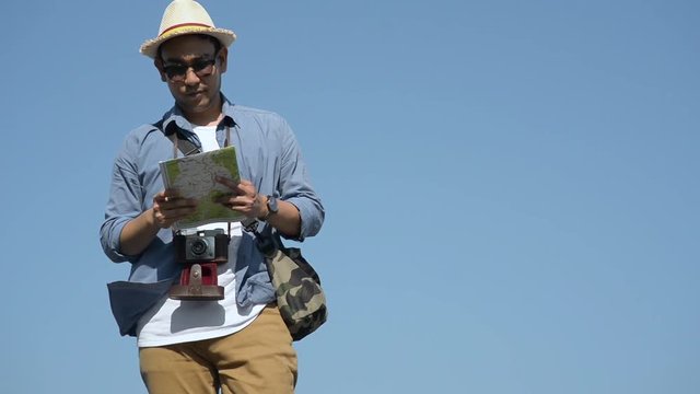 Happy Asian tourist holds map and taking pictures with vintage hipster camera while sightseeing with blue sky and sunny day.