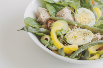 Serving of healthy salad with yellow bell pepper, eggs, tuna, green beans, spinach, arugula leaves, cherry tomato, cucumber and olives on white table