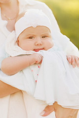 Close up cropped shot of half-year little baby girl in arms of sweet mommy. Cute child looking at camera. Soft sun light picture. Happy family spend time together in summer, white clothes