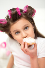 Gorgeous girl with hair curlers eating cake. Beautiful happy little girl biting on cupcake with white icing and looking at camera.