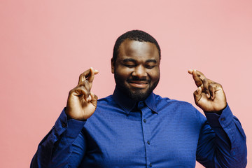 Wishing for good luck, portrait of a smiling man in blue shirt with fingers crossed, isolated on pink background