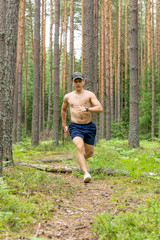 A young man with a muscular bare chest, in shorts and a cap, runs along a pine forest against the background of tree trunks