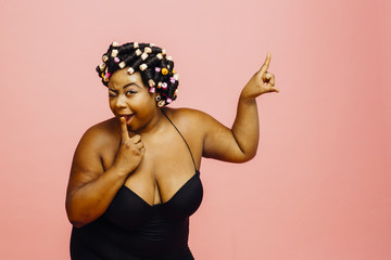 Woman with hair curlers and black camisole, winking and pointing up, isolated on pink background