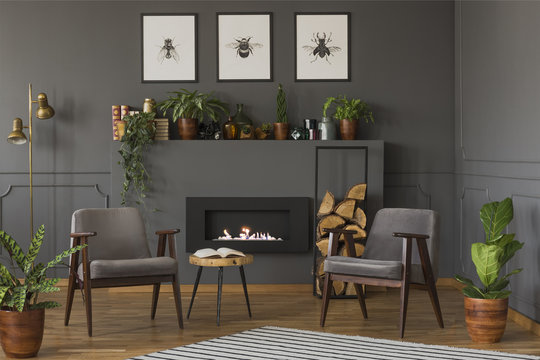 Wooden table between grey armchairs in retro flat interior with fireplace under posters. Real photo