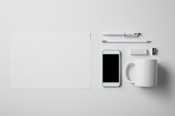 top view of smartphone with various supplies and blank paper on white tabletop for mockup