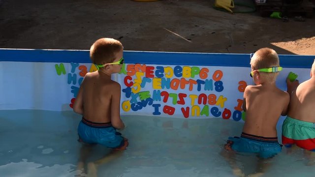 Three happy children play in swimming pool with plastic letters for bathroom. Brothers are happy together in warm water on sunny summer day. Concept of teaching games for preschoolers. Copy space text
