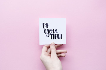 Minimal composition on a pink pastel background with girl's hand holding card with quote - be you tiful
