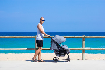 parenting and travel concept - side view of young man pushing baby stroller on the beach