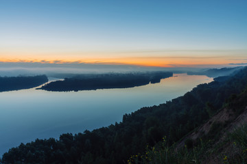 View from high shore on river. Riverbank with forest under thick fog. Dawn reflected in water. Yellow stripe in picturesque predawn sky. Colorful calm morning atmospheric landscape of majestic nature.