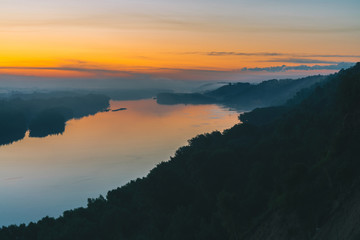 View from high shore on river. Riverbank with forest under thick fog. Gold dawn reflected in water. Yellow glow in picturesque predawn sky. Colorful morning atmospheric landscape of majestic nature.