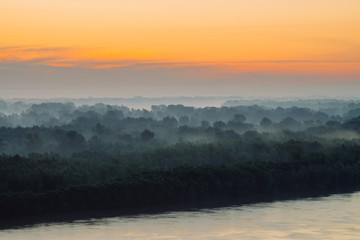 Naklejka na ściany i meble Mystical view on riverbank of large island with forest under haze at early morning. Mist among layers from tree silhouettes under warm predawn sky. Morning atmospheric landscape of majestic nature.