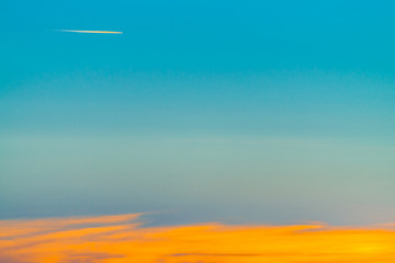 Sunrise landscape with airplane flying across blue sky and leave trail in clear sky above long orange cloudy line. Dawny cloudscape with copy space. - obrazy, fototapety, plakaty