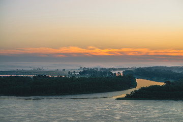 Naklejka premium Early blue sky reflected in river water. Riverbank with forest under predawn sky. Orange stripe in picturesque sky. Fog hid trees on island. Colorful morning atmospheric landscape of majestic nature.