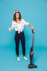 Full length portrait of outraged displeased housewife 20s doing housework and cleaning floor with vacuum cleaner, isolated over blue background