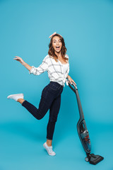 Full length portrait of joyful young woman 20s vacuuming floor with happy smile, isolated over blue background