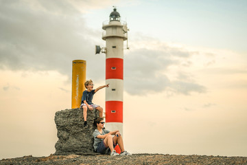 Two smiling brothers sitting near a lighthouse at the sunset with a cloudy sky in background. The youngest indicates the horizon with a finger. Two young boys with glasses sitting on the rocks