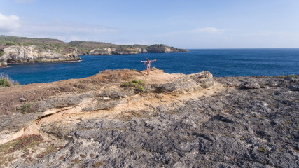 Fototapeta na wymiar Aerial view of young girl stands on the edge of a cliff and looks at the sea Nusa Penida. Girl raising her hands up on the edge of the cliff enjoys the view of the ocean. Travel concept.