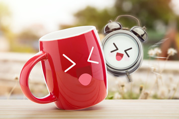 Red cup of coffee and alarm clock with a happy smile on wooden table against natural background in...