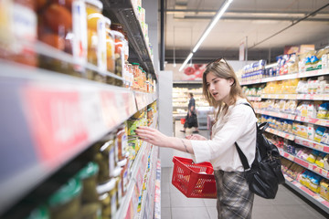 A stylish young woman with a red shopping basket in her hands selects canned vegetables in a supermarket. Attractive Girl buys canned vegetables in a supermarket