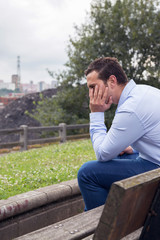Obraz premium Young male sitting on a wooden bench in a park, thinking and looking away. On a cloudy day.