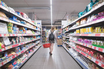 Back of a stylish woman walking in a supermarket with a basket in their hands and buying products. Shopping in a supermarket. Girl walks a supermarket and chooses shopping
