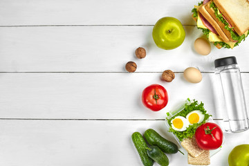 Fototapeta na wymiar Lunch. Sandwich and fresh vegetables, bottle of water, nuts and fruits on white wooden background. Healthy eating concept. Top view with copy space