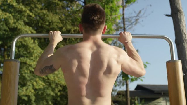 Slow motion, close up unrecognizable fit tattooed man doing pull ups on a sunny day in green park with outdoor gym equipment