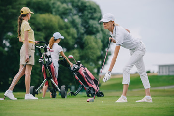Selective focus of focused woman in cap playing golf with friends near by at golf course