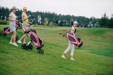 Fototapeta premium Side view of women in polos and caps with golf gear walking on green lawn at golf course